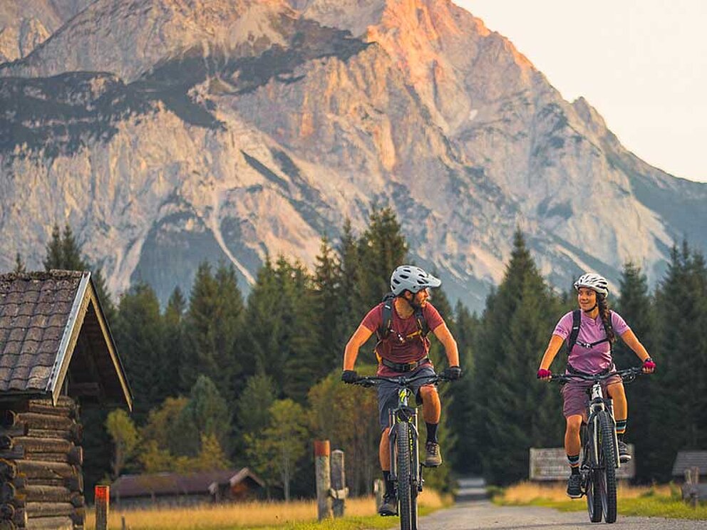 2 E-Mountainbiker in flotter Fahrt auf einer einsamen Straße. Dahinter eine felsige Bergwand.