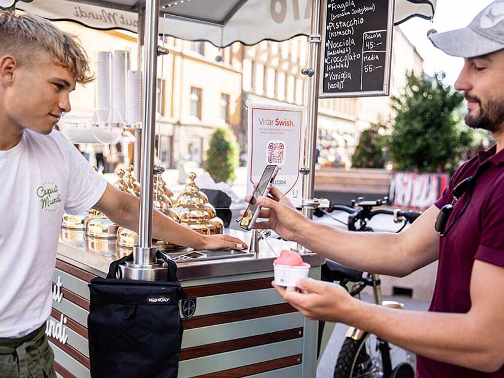 ein Mann mit Kappe kauft Eis bei einem Fahrrad-Eisverkäufer in Schweden. Er bezahlt bargeldlos mit Mobiltelefon.