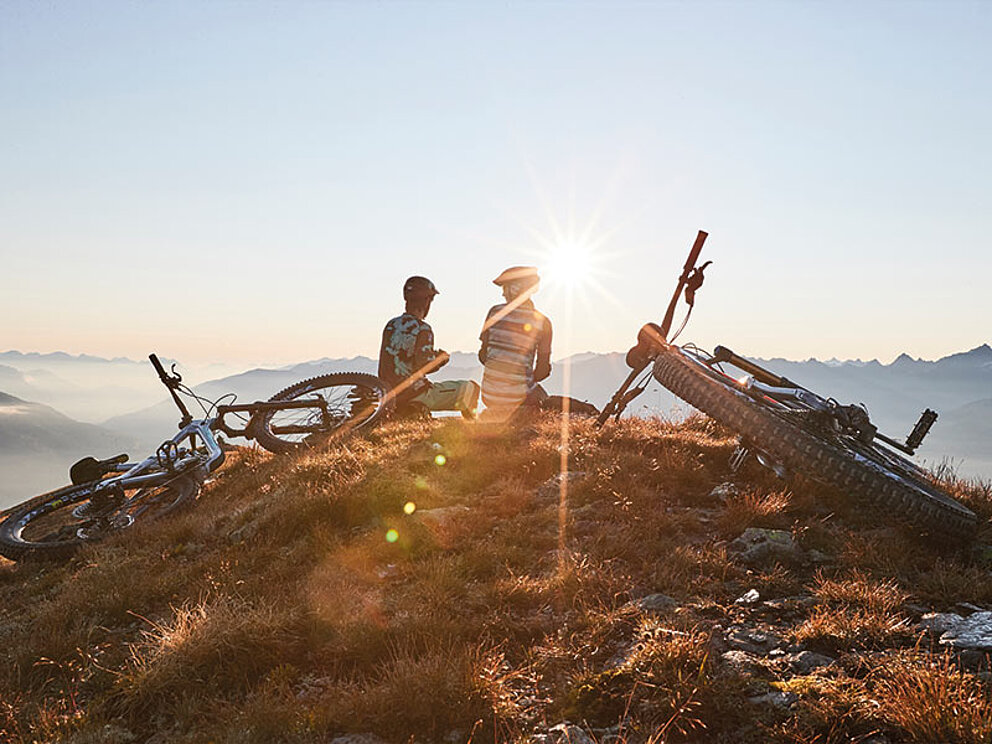2 Biker genießen die Aussicht im Sonnenuntergang. Die 2 Bikes liegen neben den Personen.