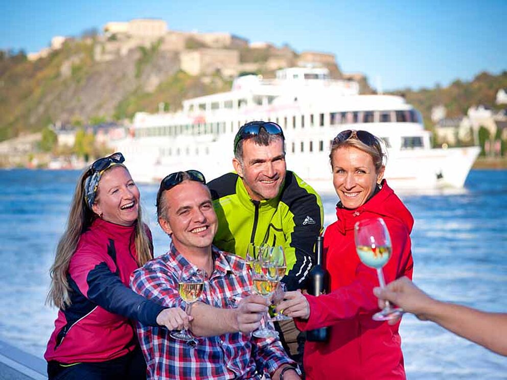 Gruppe Radfahrer beim Weintrinken am Rheinufer in Koblenz mit Schiff vor Festung