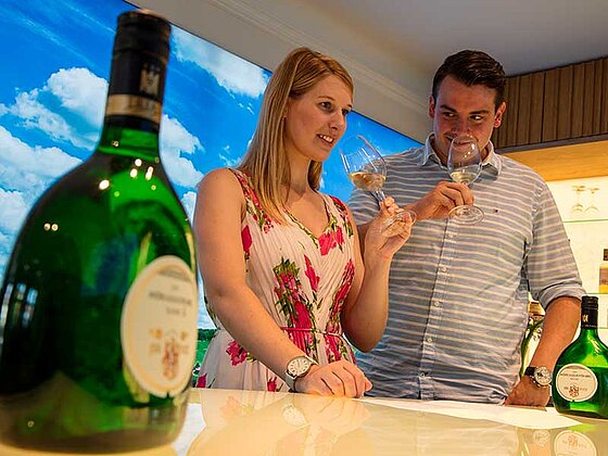 Mann und Frau verkosten Frankenwein. Sie halten Weingläser in den Händen, Bocksbeutelflaschen stehen davor.