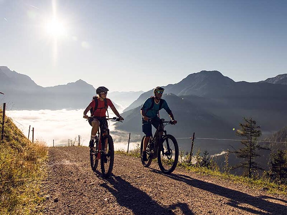 2 Mountainbiker im sonnigen Hochgebirge. Über dem liegt eine dicke Nebeldecke