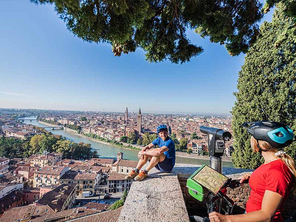 ein Mann sitzt auf einer Mauer und blickt über die unter ihm liegende Stadt, eine Frau mit Ihrem Rad steht im Vordergrund