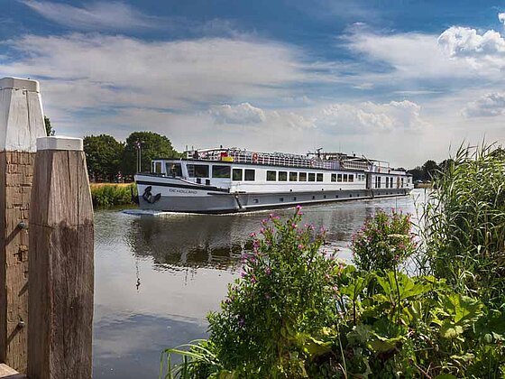 Flussschiff De Holland auf einem flachen Fluss. Im Vordergrund Pfeiler einer Anlegestelle un Büsche