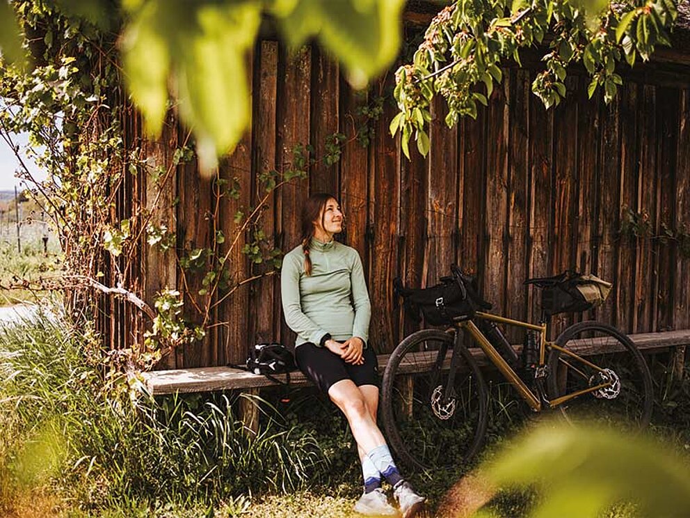 1 Dame im Bike-Outfit sitzt auf einer Bank. Neben ihr ihr Gravelbike und Obstbäume.