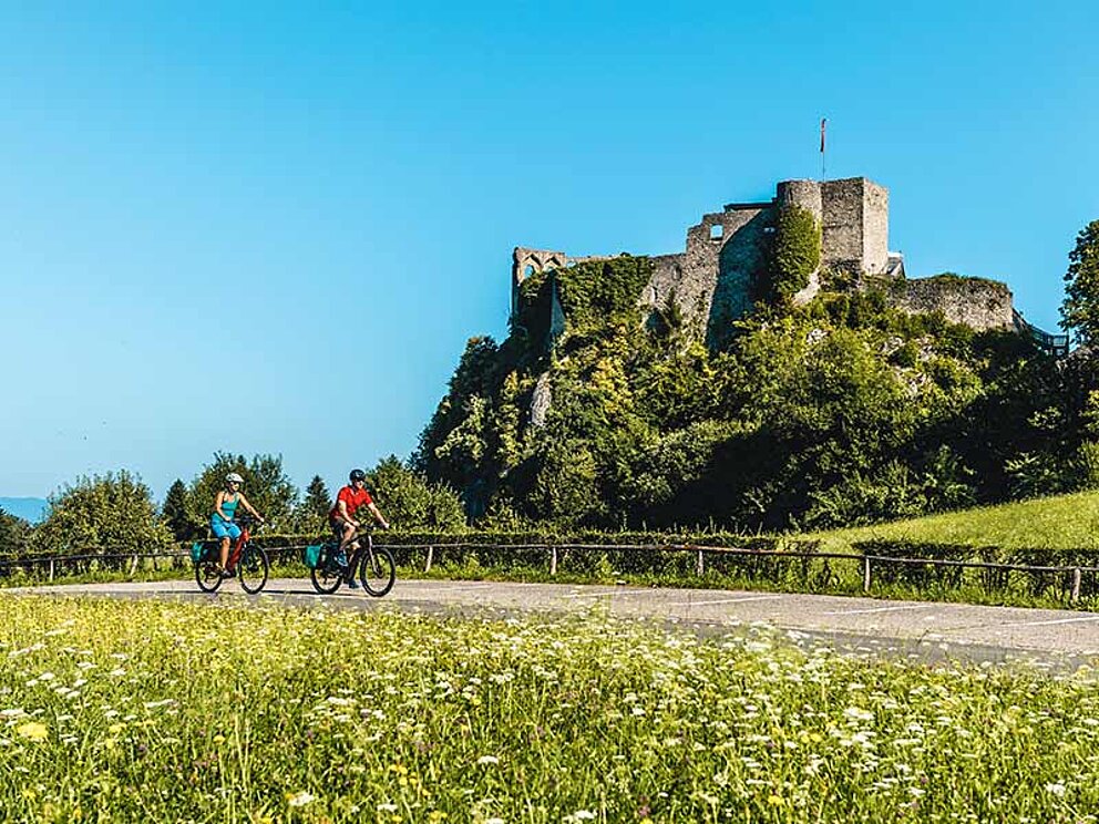 2 Radfahrer vor der Burg Finkenstein, im Hintergrund der Faaker See