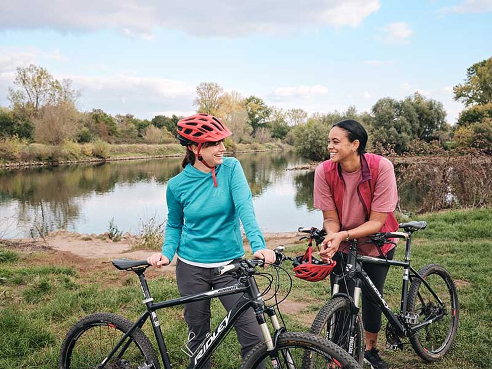 2wei Frauen stehen in der Aulandschaft des Rheins und unterhalten sich. Sie lehnen sich an ihre Frahrräder