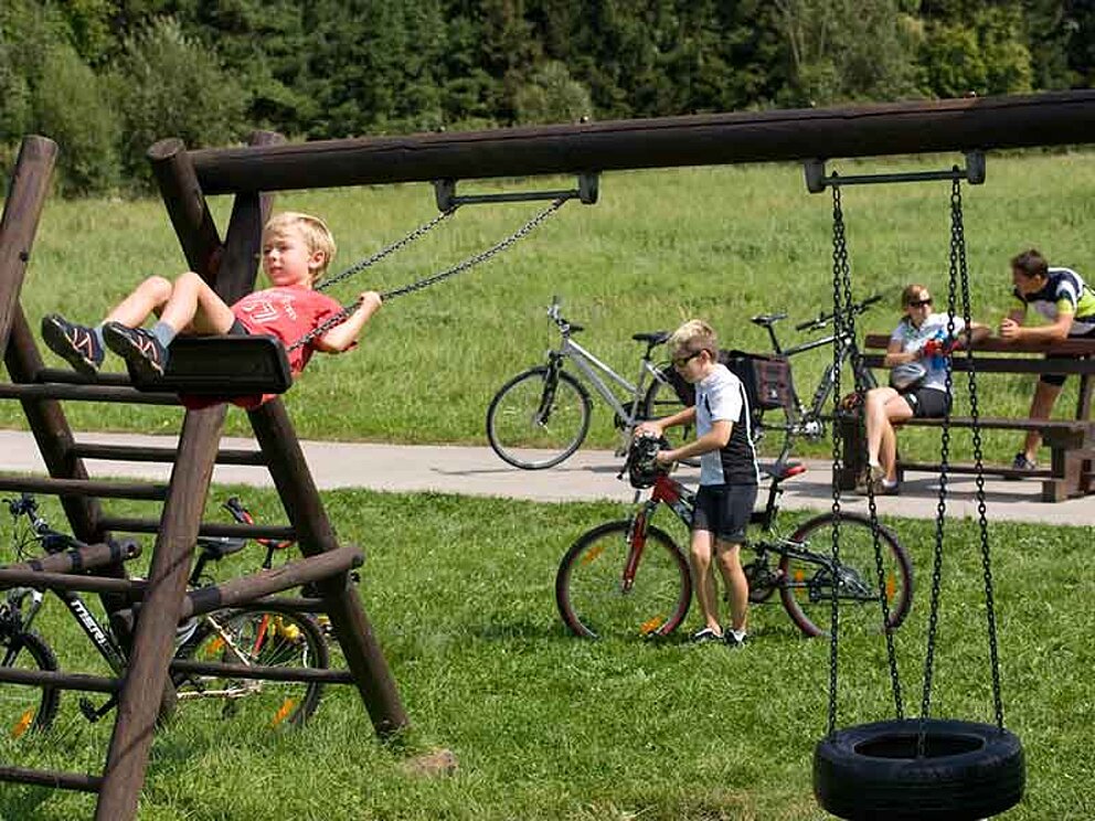 Kleiner Bub schaukelt, großer Bub wartet auf dem Fahrrad, die Eltern sitzen auf einer Bank