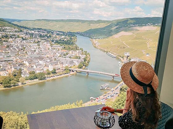 Eine Dame mit Hut sitzt am Panoramafenster auf der Burg Landshut und blickt über die Stadt Bernkastel-Kues und die Mosel.