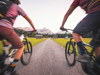 2 Biker flitzen über eine einsame Straße. Links und rechts Wiesen, in der Ferne Berge.