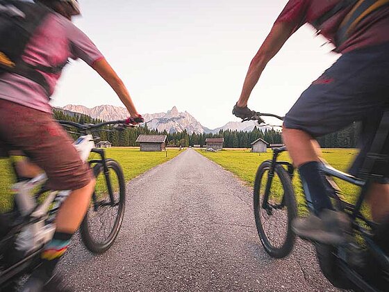 2 Biker flitzen über eine einsame Straße. Links und rechts Wiesen, in der Ferne Berge.