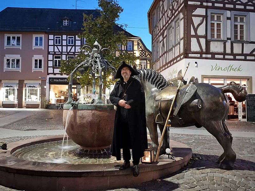Ein Nachtwächter mit Umhang, Hut und Hellebarde lehnt an einem Brunnen mit Pferdeskulptur. Dahinter Fachwerkhäuser