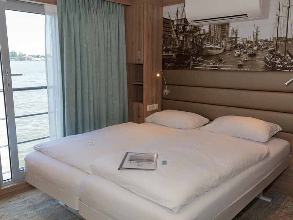 Ein großes Doppelbett in der Kabine. Dahinter ein Wandbild mit Hafen, links davon ein großes Schiebefenster