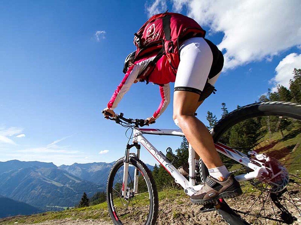 Frau stehend am Mountainbike von hinten fotografiert. Im Hintergrund alpine Bergwelt