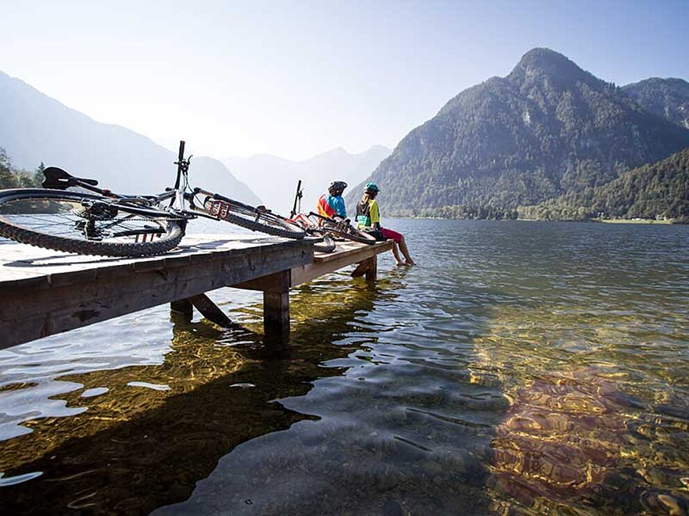 2 Personen sitzen auf einem Steg am Gosausee und lassen die Füße ins Wasser hängen. Am Steg liegt ein Mountainbike.