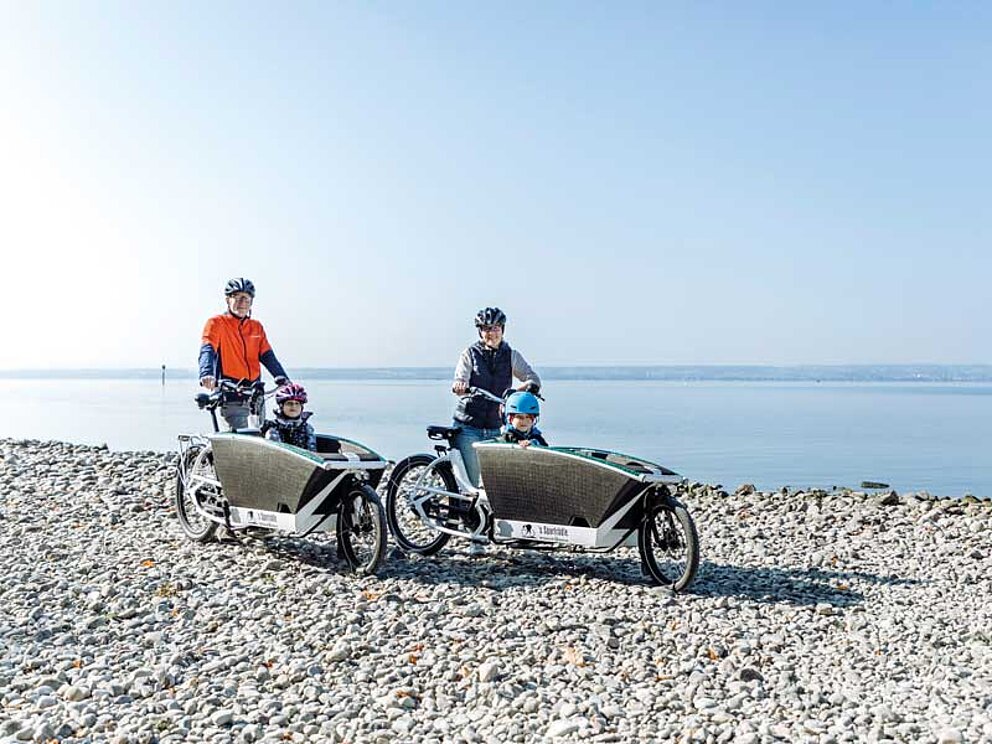 Mann und Frau mit Lastenfahrrad in denen Kinder sitzen, direkt am Bodenseeufer