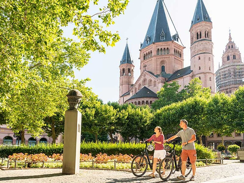 Eine Dame und ein Mann schieben ihre Fahrräder durch einen Park. Dahinter der Wormser Dom