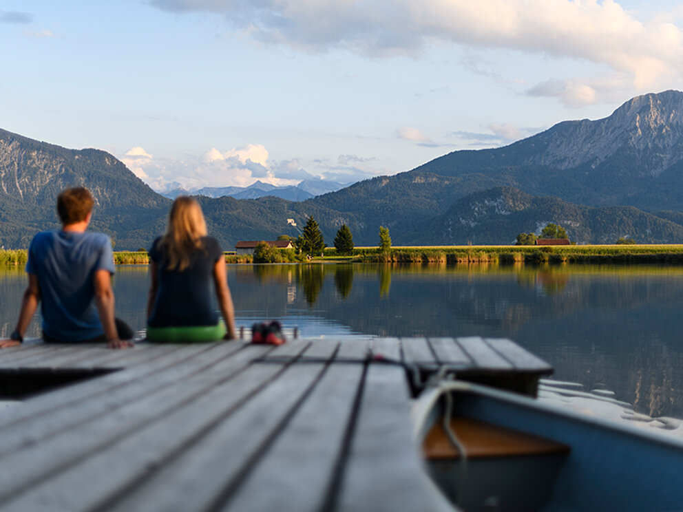 Ein Mann und eine Frau sitzen am Steg und lassen die Füße in den Staffelsee baumeln.