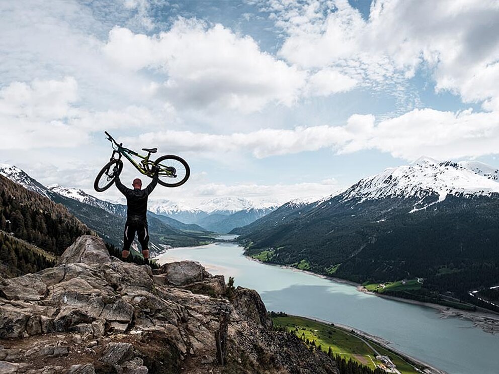 Ein Mountainbiker hebt sein Bike in die LUft, im Hintergrund ein Bergsee