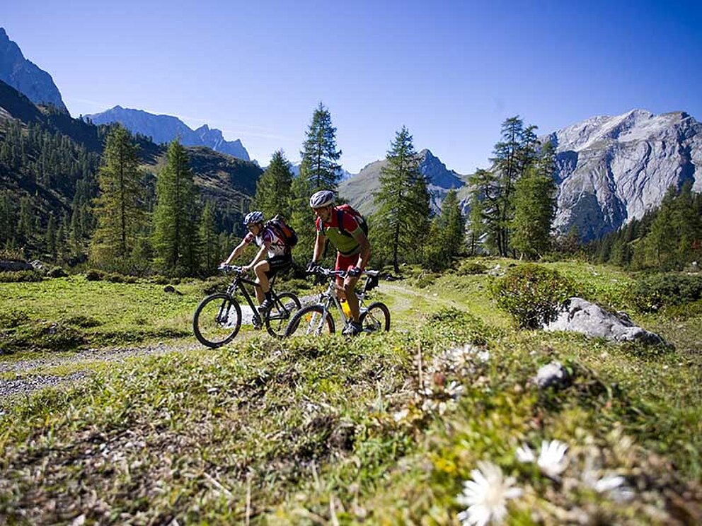 2 Mountainbiker fahren hintereinander auf einem steinigen Gebirgsweg. Im Hintergrund alpine Bergwelt.