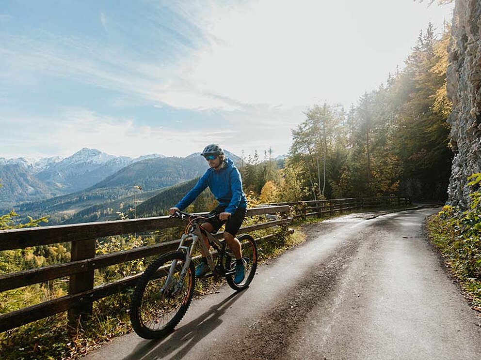 Ein Mountainbiker am Schotterweg, im Hintergrund ein Geländer und Bergpanorama