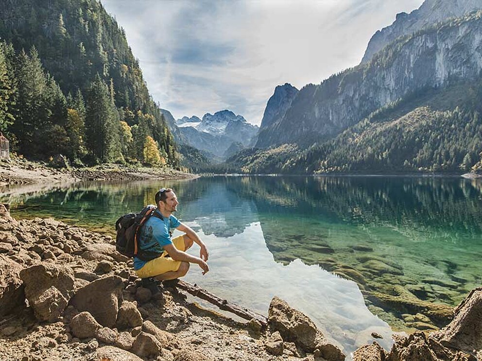 ein Man kniet am Ufer des Gosausees, im Hintergrund der Dachstein