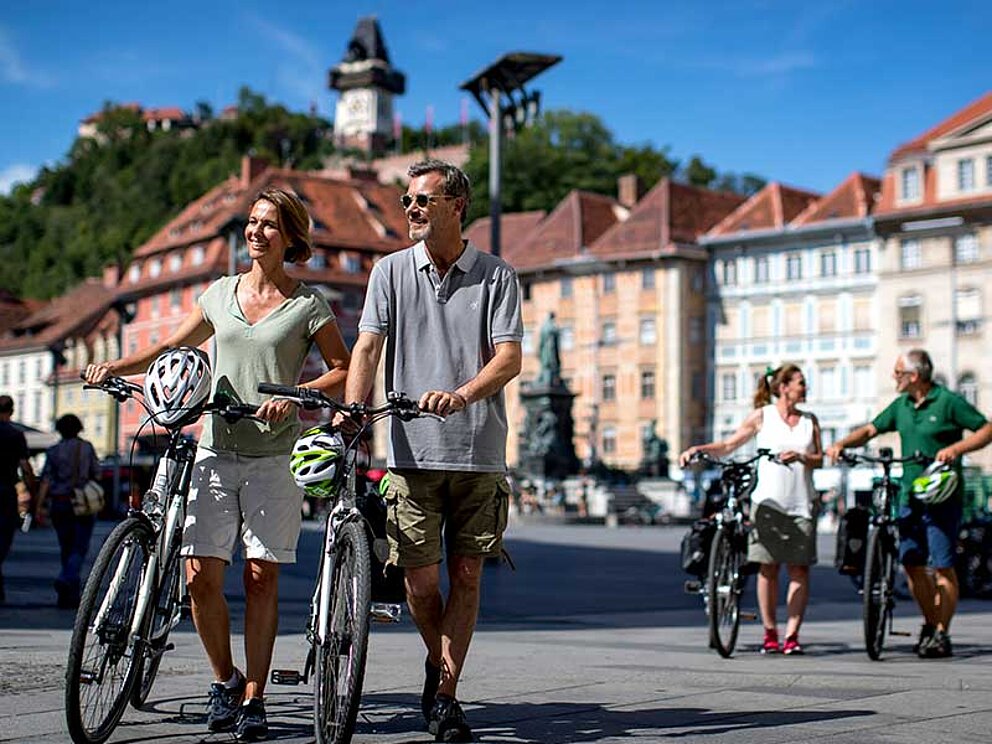 Ein Mann und eine Frau schieben ihre Fahrräder über den Hauptplatz in Graz.