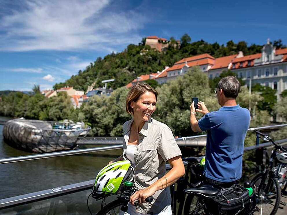Ein Mann und eine Frau stehen am Mursteg in Graz. Der Mann fotgrafiert den Fluss Mur unterhalb.