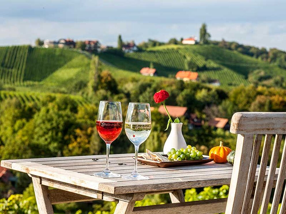 Rot- und Weißwein, sowie Weintrauben stehen auf einem Holztisch. Davor befindet sich ein Holzstuhl. Im Hintergrund die malerischen Hügel der Südsteirischen Weinstraße.