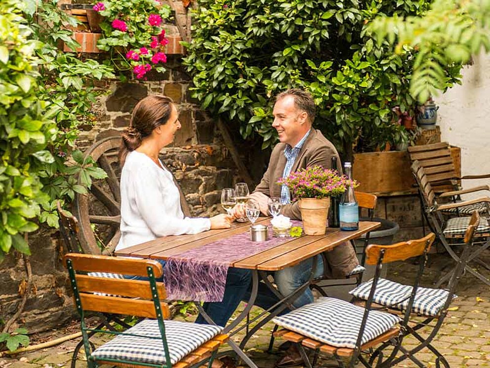 ein Paar sitzt an einem Tisch im Gastgarten einer Weinschenke, vor ihnen jeweils ein Glas Weißwein.