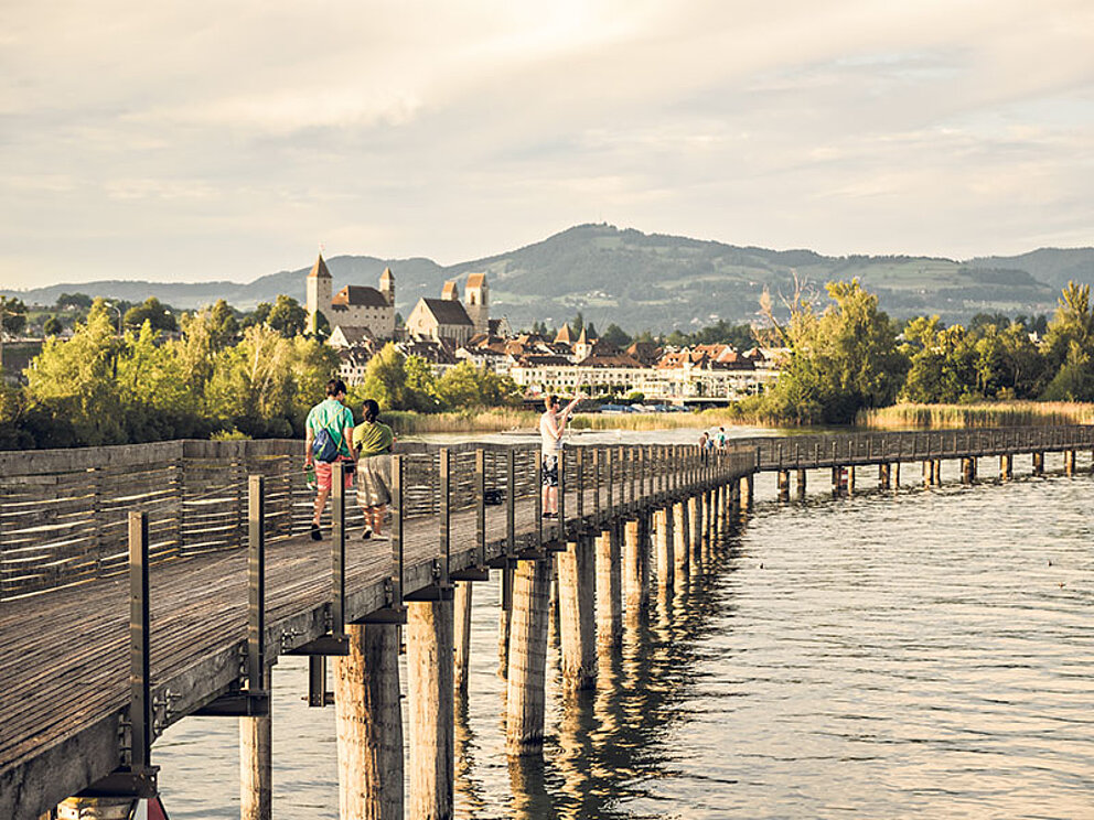 2 Personen spazieren über einen Holzsteg am Zürichsee.