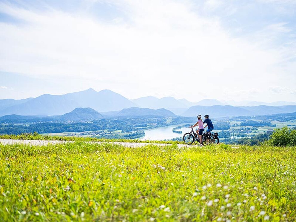2 Radfahrer, im Vordergrund eine Blumenwiesen, im Hintergrund der See und Bergpanorama