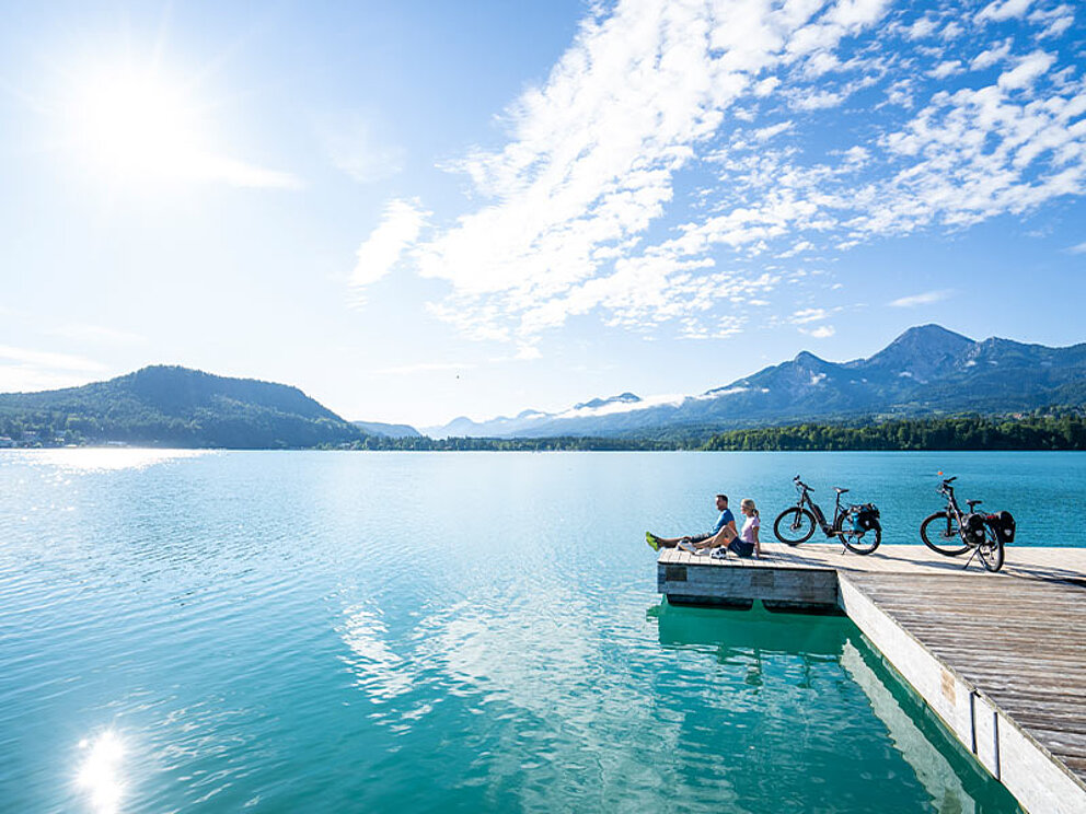 2 Radfahrer sitzen am Steg und schauen über den See, im Hintergrund Berge