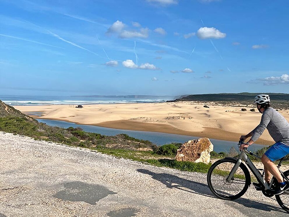 ein Radfahrer am Schotterweg, im Hintergrund die Küste mit Sandstrand