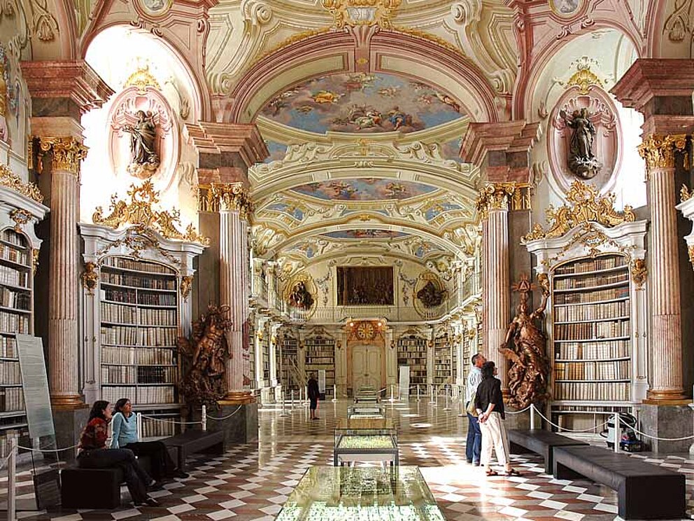 Die Bibliothek im Stift Admont. Die Bibliothek ist mit üppigen Deckengemälden und viel Stuck an den Wänden ausgestattet. Englang der Wände sind hohe Bücherregale. Menschen bewundern die Pracht.