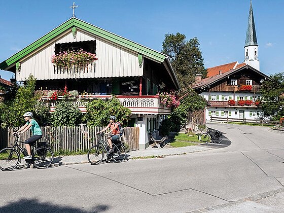 2 Radfahrer sausen an typisch oberbayerischen Häusern vorbei.