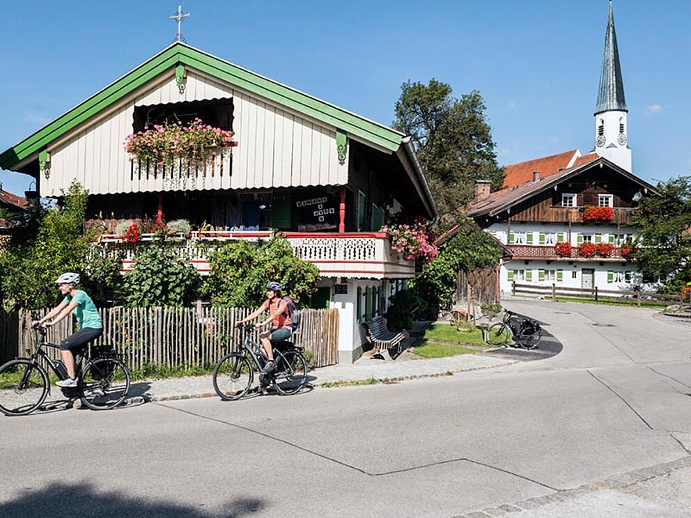 2 Radfahrer sausen an typisch oberbayerischen Häusern vorbei.