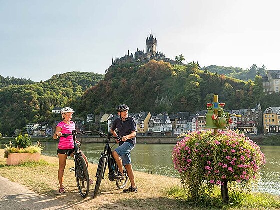 2 Radfahrer stehen am Ufer der Mosel, im Hintergrund die Reichsburg und Weinberge bei Cochem