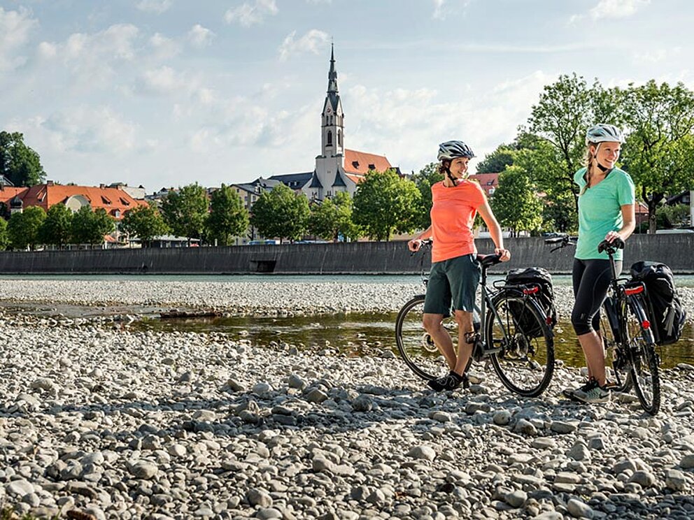 2 Radfahrer auf einer Schotterbank, im Hintergrund der Kirchturm von Bad Tölz.