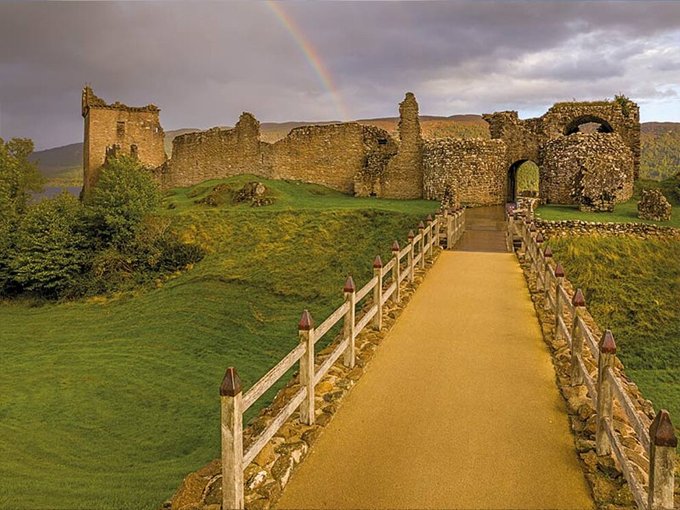 Die Ruine von Urquhart Castle in der Abenddämmerung, im Hintergrund ein Regenbogen.
