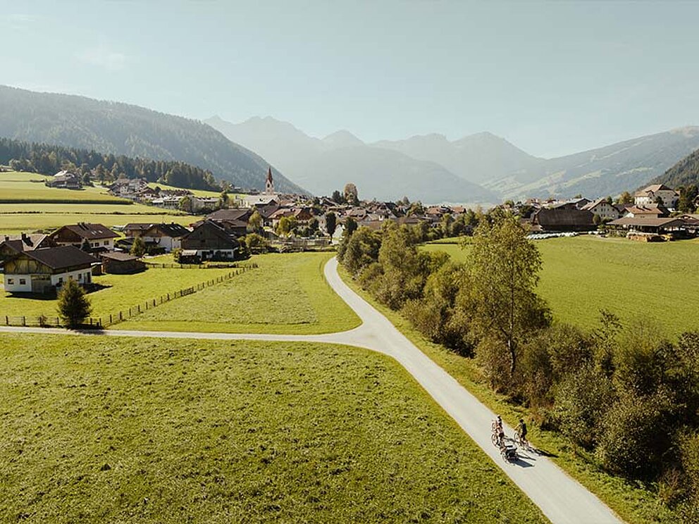 2 Radfahrer am asphaltierten Radweg von oben fotografiert, im Hintergrund ein Ort und Gebirge