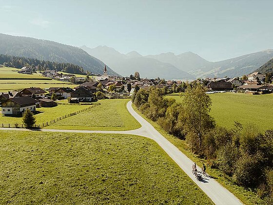 2 Radfahrer am asphaltierten Radweg von oben fotografiert, im Hintergrund ein Ort und Gebirge