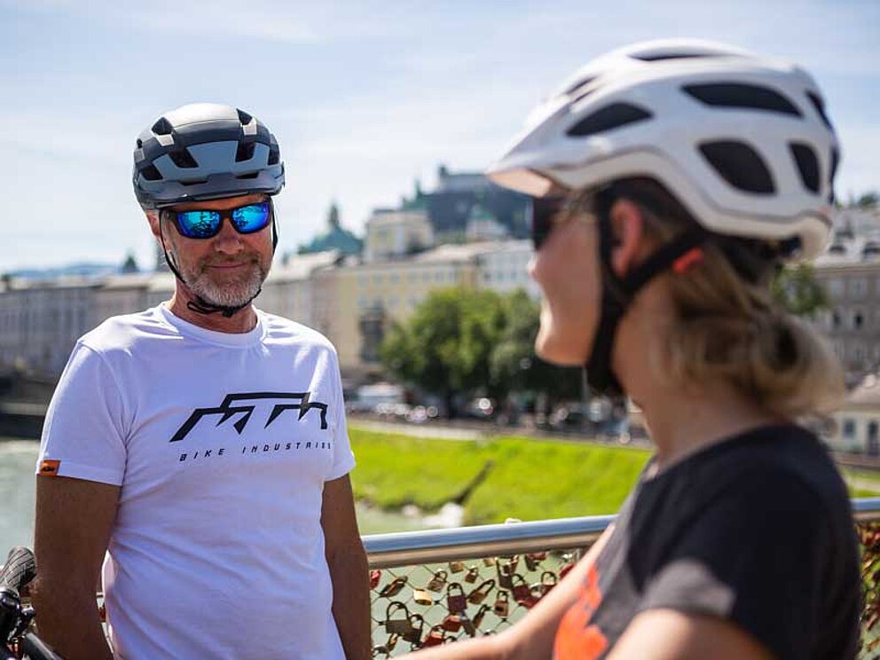 Mann und Frau mit Radhelm und Raddress in Großaufnahme, dahinter die Festung Hohensalzburg