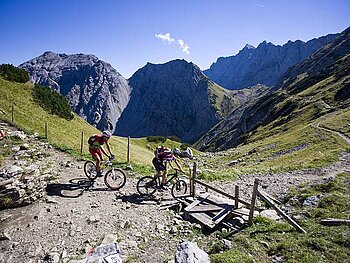 2 Mountainbiker fahren hintereinander auf einem steinigen Gebirgsweg. Im Hintergrund alpine Bergwelt.