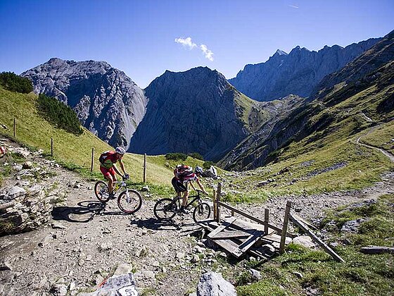 2 Mountainbiker fahren hintereinander auf einem steinigen Gebirgsweg. Im Hintergrund alpine Bergwelt.