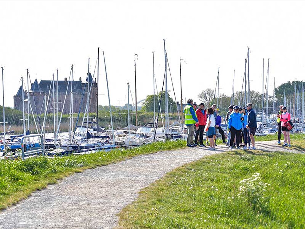 Radfahrer pausieren im Hafen von Mudlen, im Hintergrund Segelschiffe im Hafen liegend.