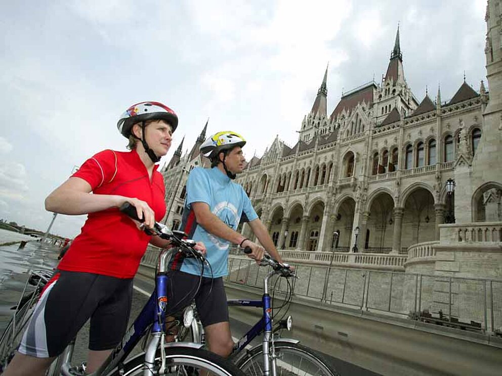 Ein Mann und eine Frau in Raddressen stehen mit ihren Fahrrädern vor dem Parlament in Budapest. Das Gebäude hat eine neugotische Fassade mit aufstrebenden Türmchen und Fenstern. Im Erdgeschoß dominiert ein Bogengang.