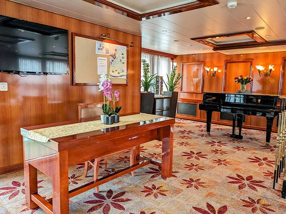 Empfangsbereicht mit Teppichboden, einer Rezeption mit Flatscreen an der Wand, im Hintergrund ein Flügel