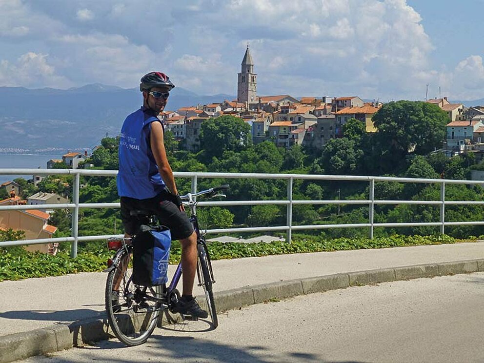 Ein Radfahrer auf einer Straße, im Hintergrund der Ort Vrbnik mit hohem Kirchturm