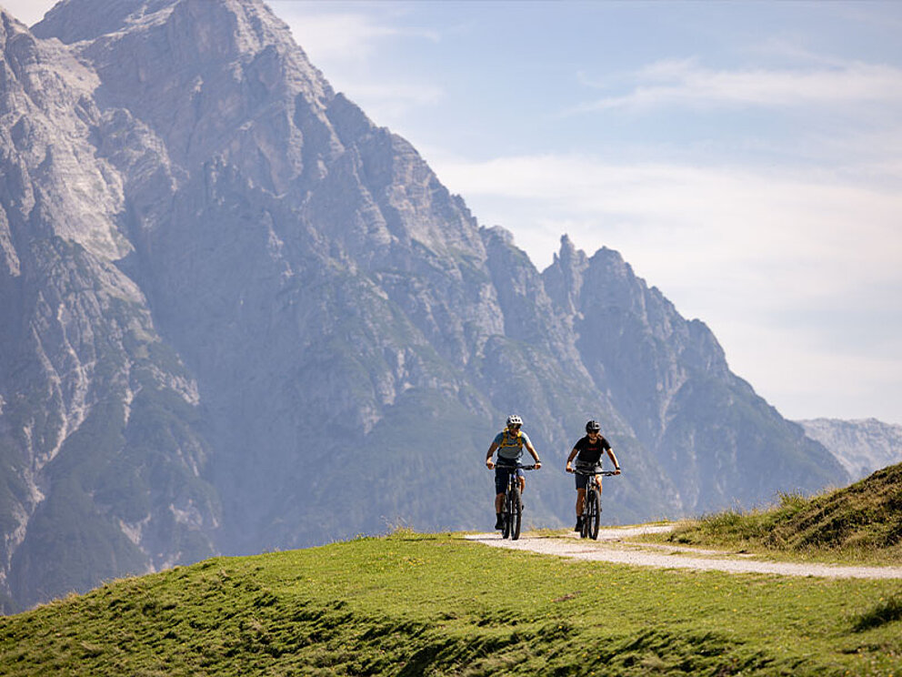 2 Biker auf einem Schotterweg, im Hintergrund beeindruckende Berge